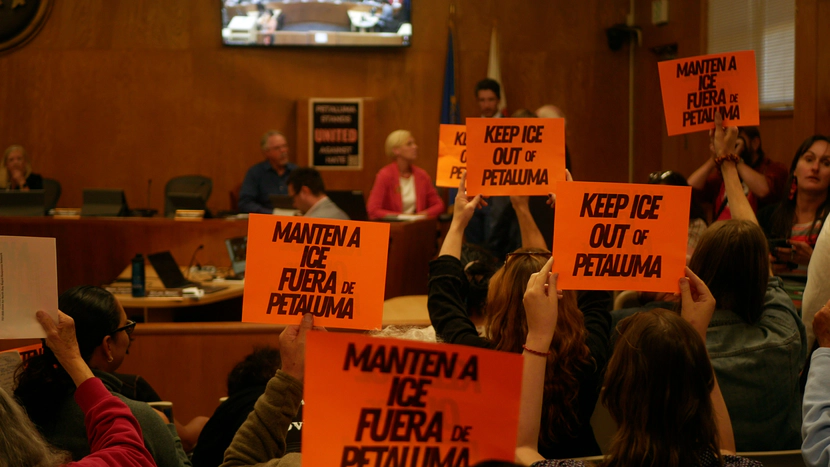 Anti- ICE community members inside the Petaluma City Hall holding orange signs that say KEEP ICE OUT OF PETALUMA and MANTEN A ICE FUERA de PETALUMA