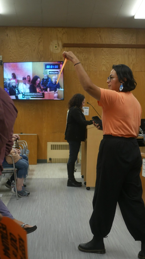 Woman in orange shirt holds a sign up inside Petaluma City Hall meeting